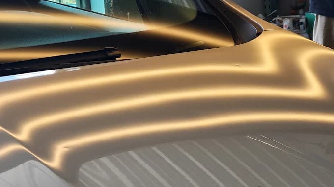 Close-up under reflective light board revealing hail dents across a vehicle hood and windshield cowl